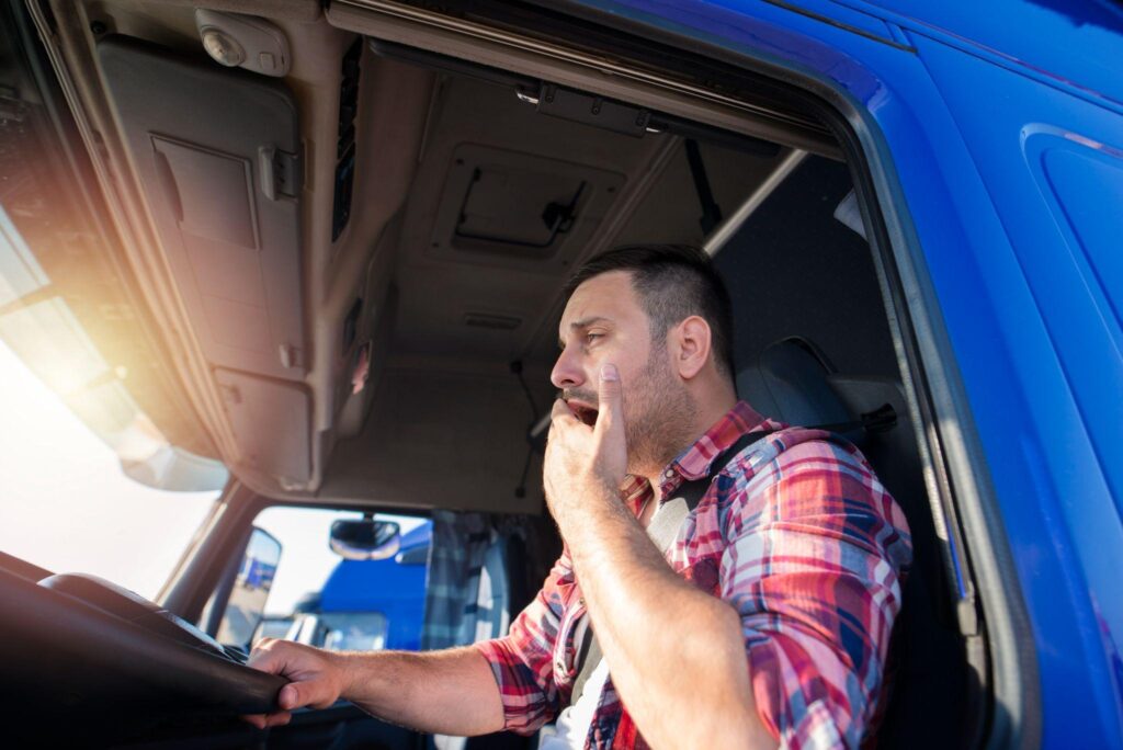 Man wearing red plaid shirt yawning while behind steering wheel of semi rig