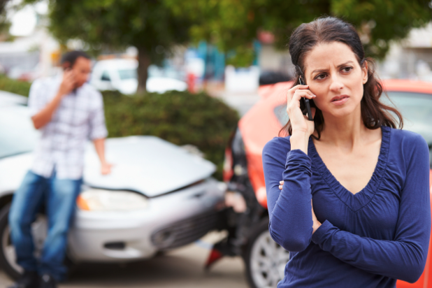 Two wrecked vehicles in blurry background with man on phone, woman on phone in foreground. 